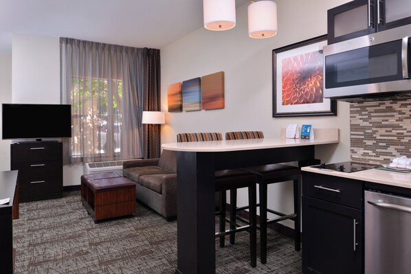 Habitación de hotel compacta con kitchenette, televisor de pantalla plana, sofá, comedor y un ventanal grande con cortinas.