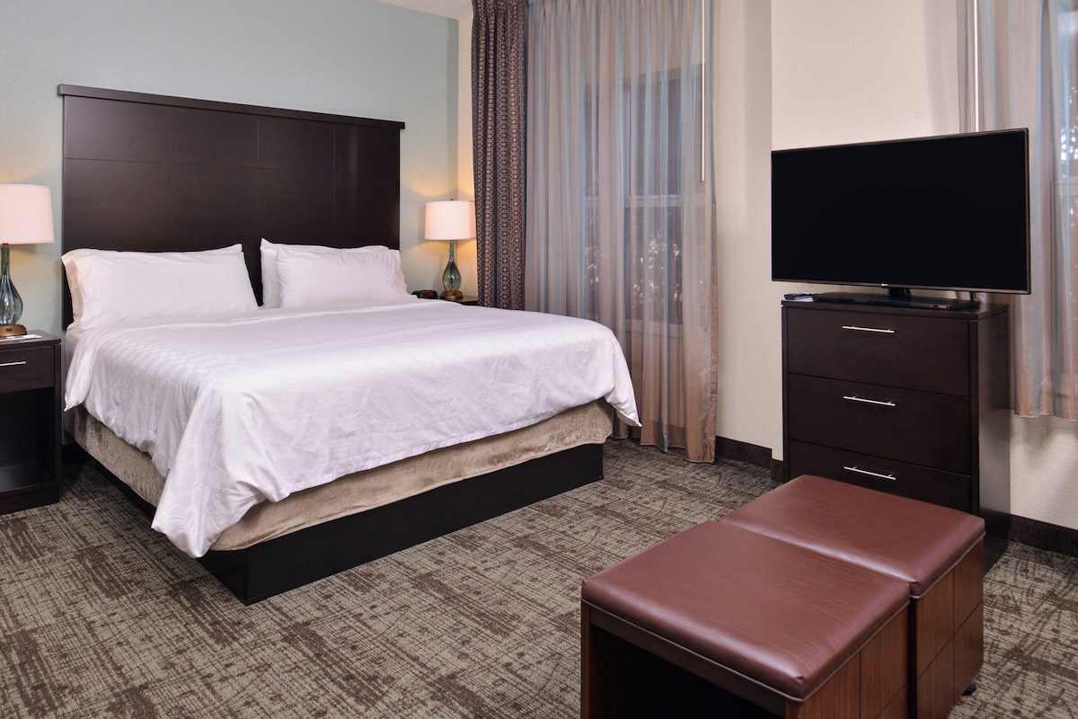 Quarto de hotel com cama grande, televisão de tela plana, escrivaninha com luminária e um banco.