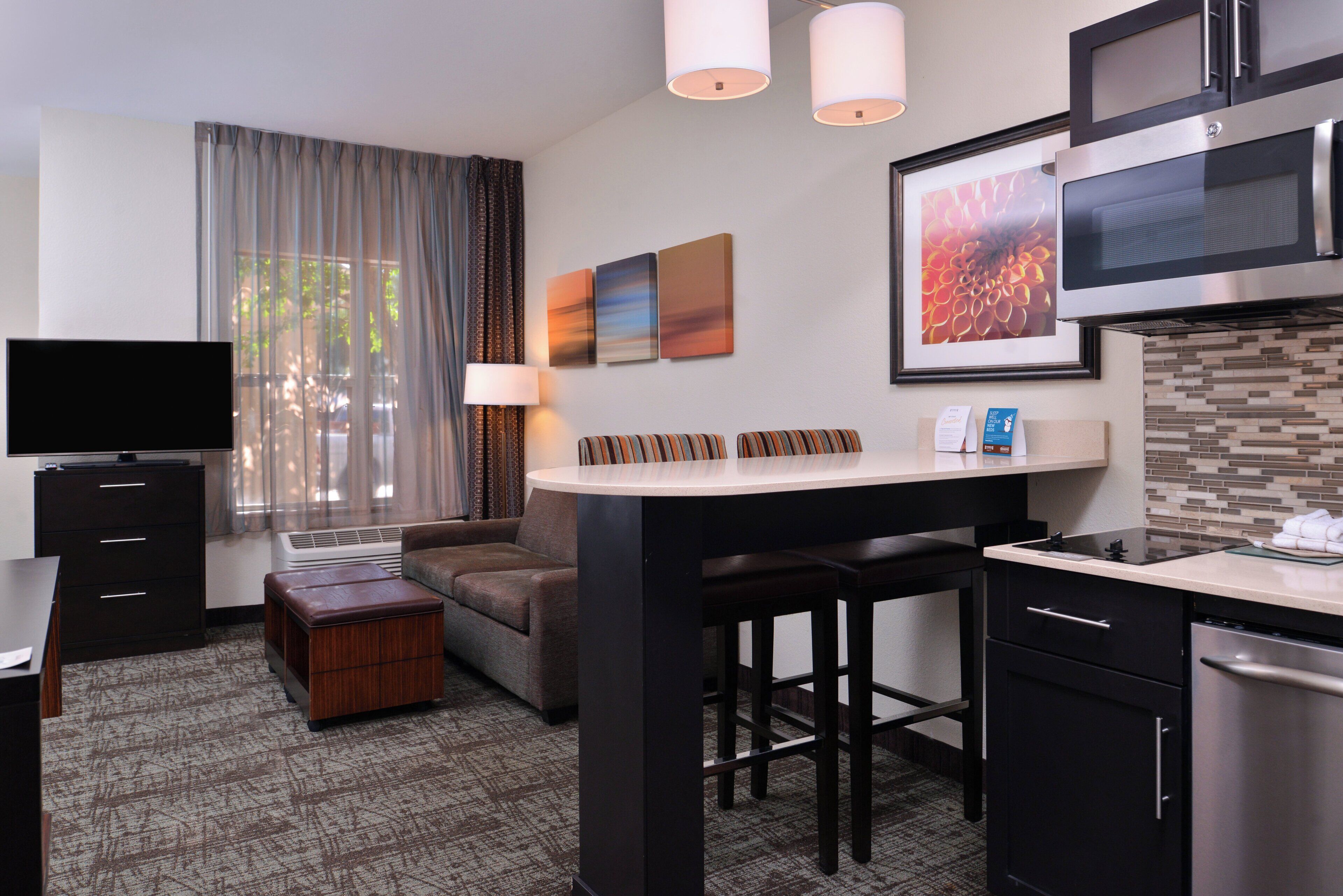A compact hotel room with a kitchenette, a flat-screen TV, a sofa, a small table, and a bar area.