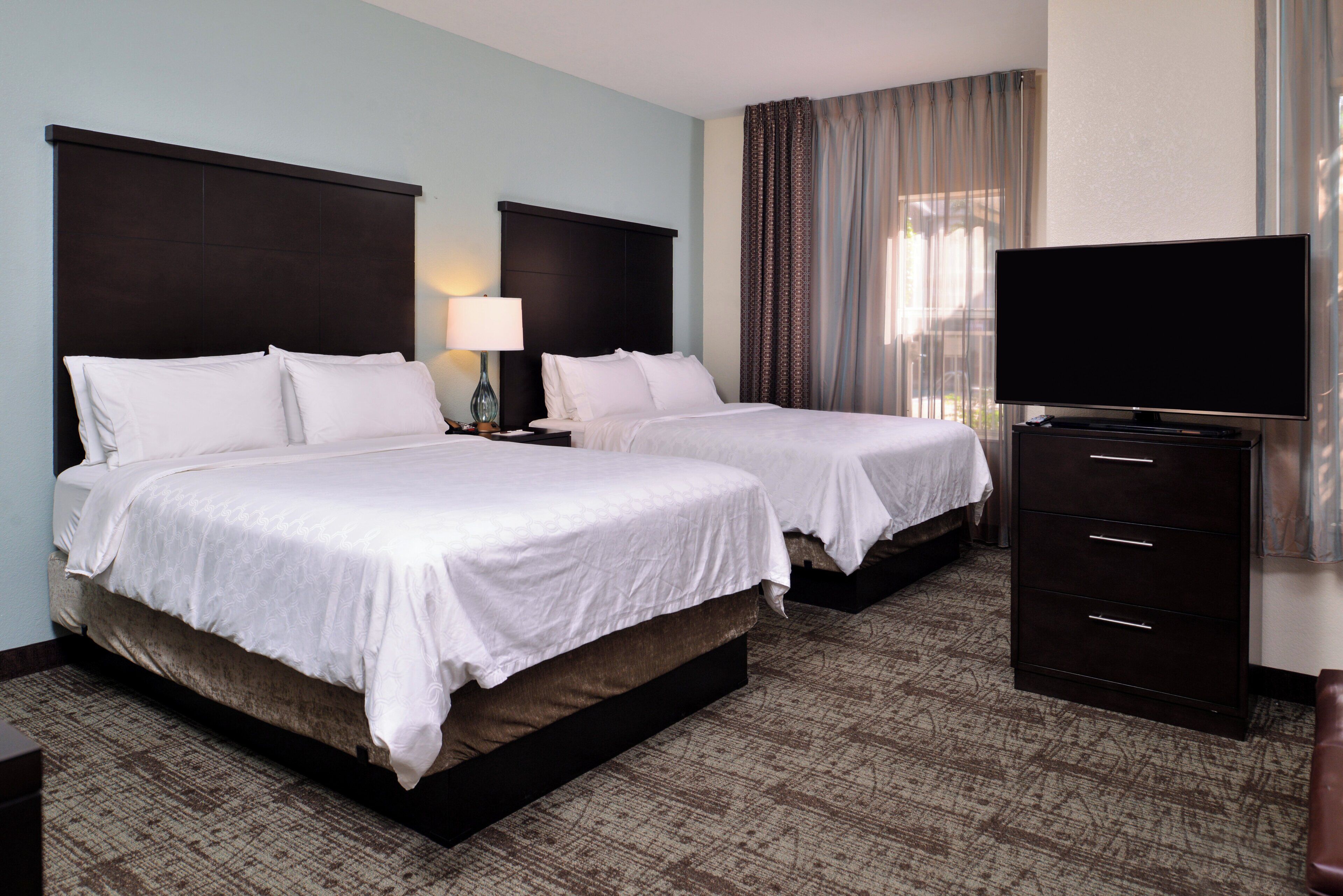 A hotel room with two beds, a flat-screen TV, a dresser, and a window with curtains.