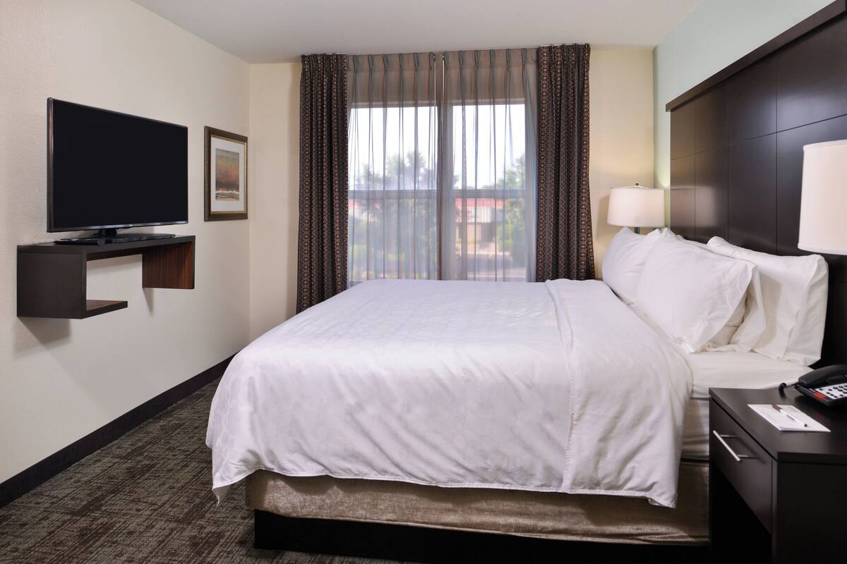 Een hotelkamer met een groot bed, een flatscreen televisie aan de muur, een nachtkastje met een lamp en een raam met gordijnen.