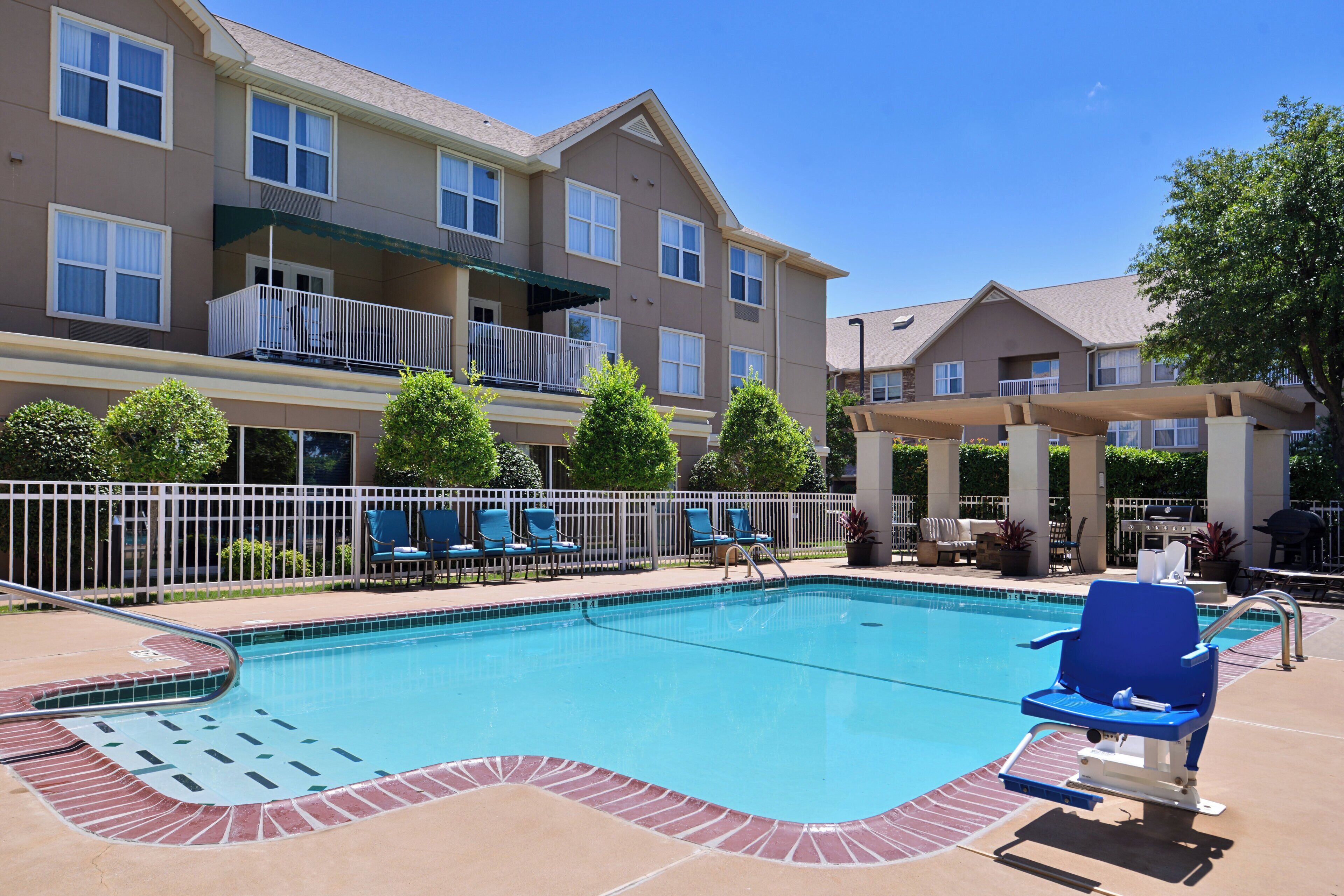 A swimming pool with lounge chairs and a barbecue area, surrounded by a fence and a multi-story building with balconies.
