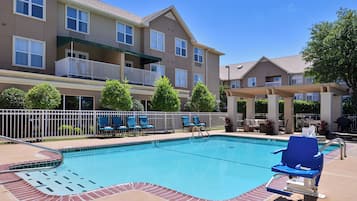 A swimming pool with lounge chairs and a barbecue area, surrounded by a fence and a multi-story building with balconies.