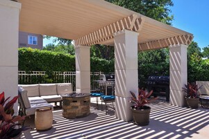 Un patio exterior con pérgola, mobiliario, una parrilla y plantas en macetas.