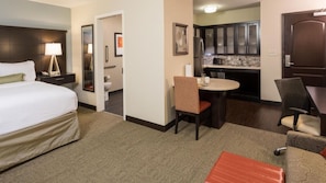 Quarto de hotel com cama, escrivaninha, cadeira, mesinha e cozinha compacta.