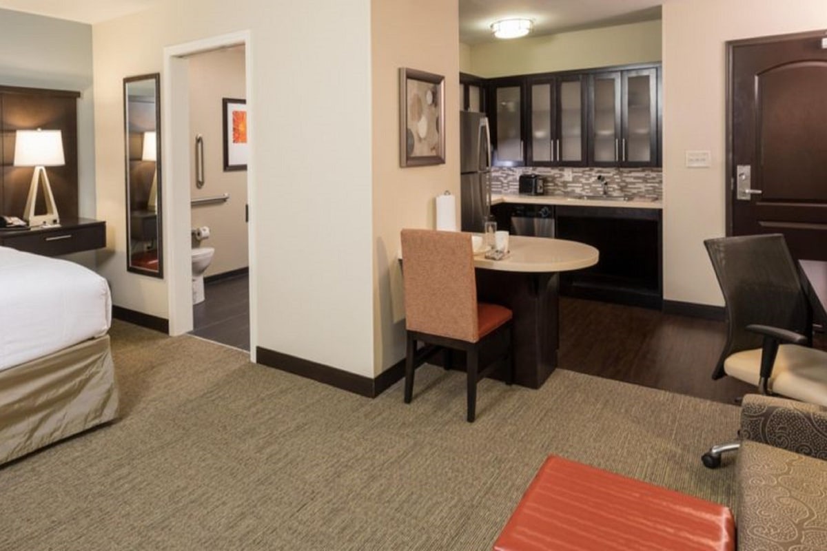 Quarto de hotel com cama, escrivaninha, cadeira, mesa pequena, cozinha compacta e banheiro.