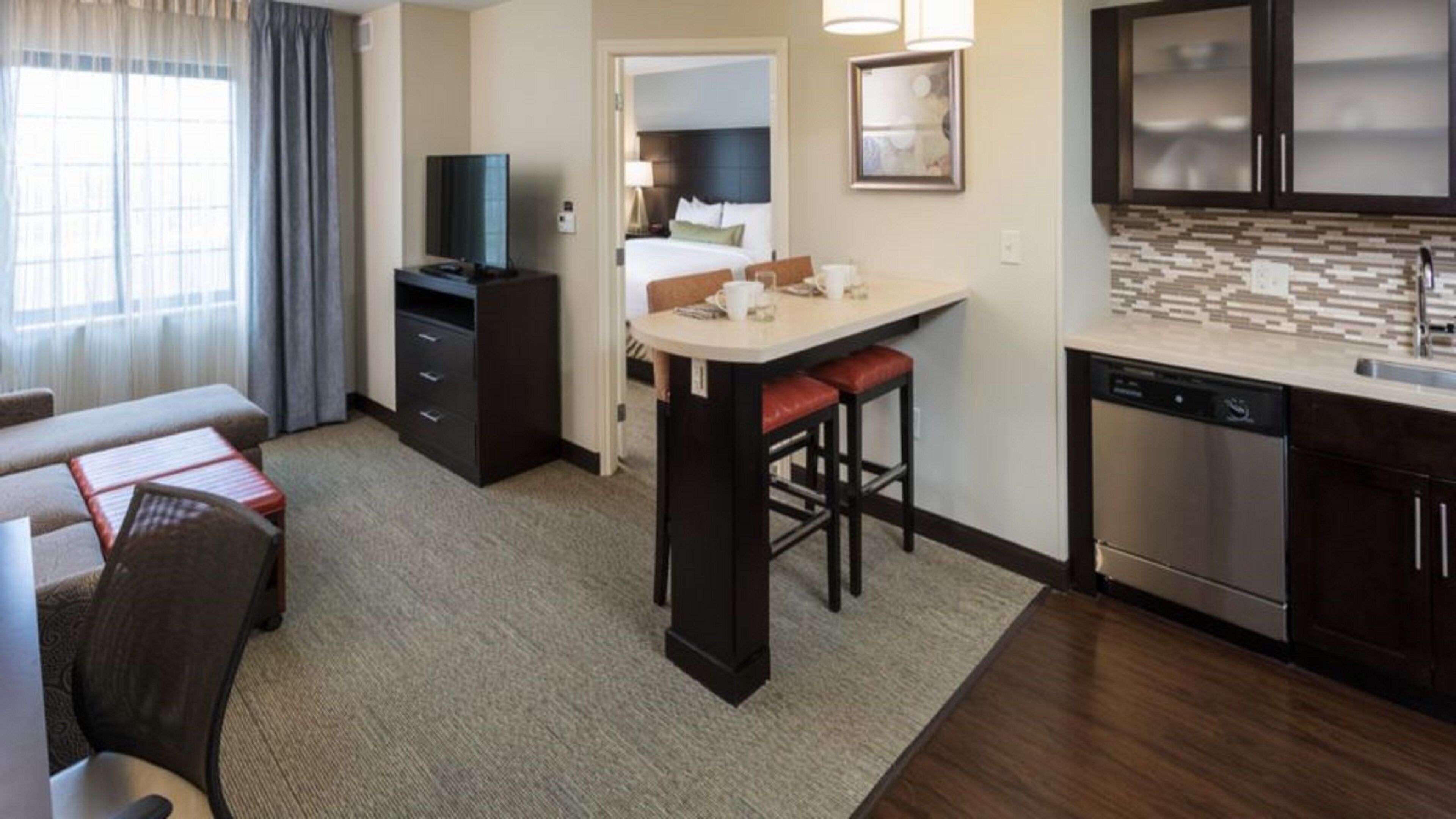A hotel room with a kitchenette, a dining area, a living room with a sofa and armchair, and a large window with curtains.