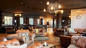 A spacious lounge area with leather sofas and armchairs, a bar in the background, and large windows allowing natural light.