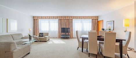 A hotel room with a sofa, a TV, a dining table with chairs, and a large window with curtains.