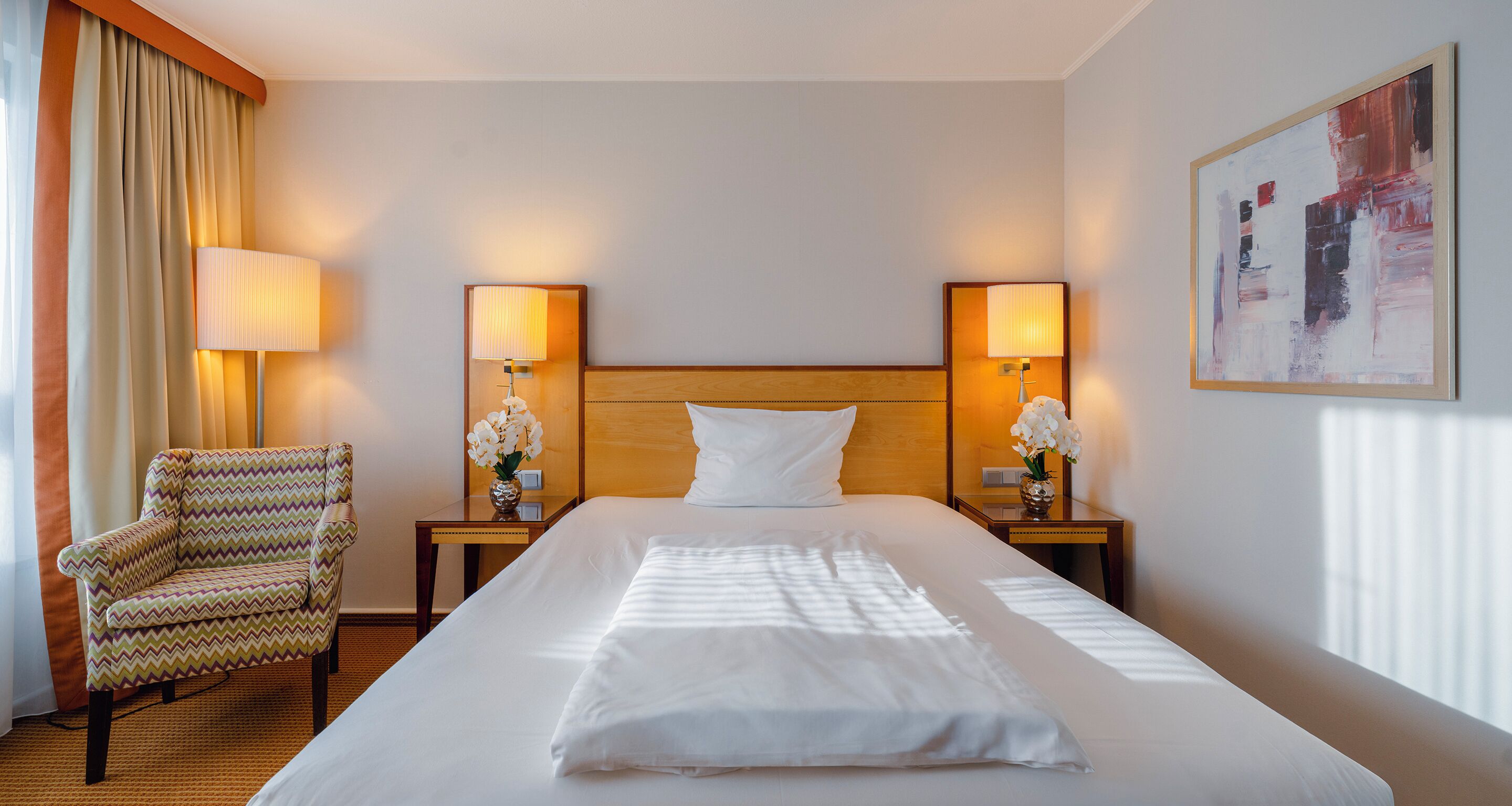 A hotel room with a large bed, two bedside lamps, a chair, and a painting on the wall.