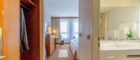 A hotel room with a bed, a desk, a chair, a bathroom with a sink and mirror, and a closet with clothes hanging.