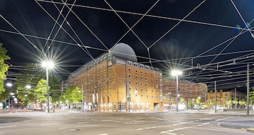 Un edificio moderno con una cúpula destacada y varios pisos, rodeado por una calle urbana bien iluminada de noche.