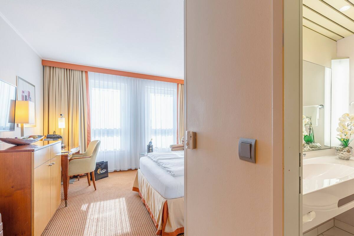 Een hotelkamer met een bed, een bureau, een stoel, een bureaulamp, een raam met gordijnen en een badkamer met een wastafel en spiegel.
