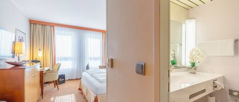 A hotel room with a bed, a desk, a chair, a desk lamp, a window with curtains, and a bathroom with a sink and mirror.