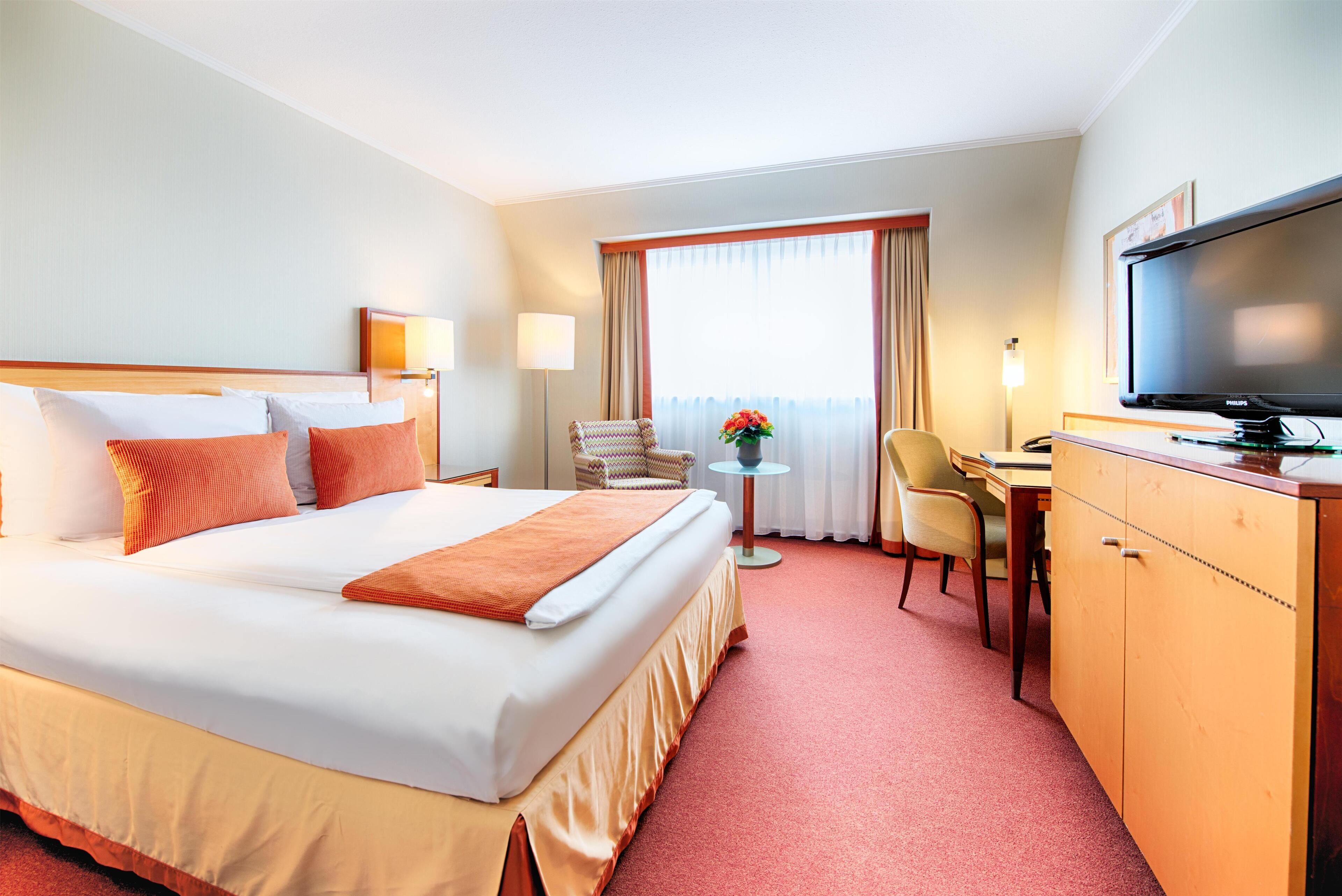 A hotel room with a large bed, a desk, a chair, a television, and a window with curtains.