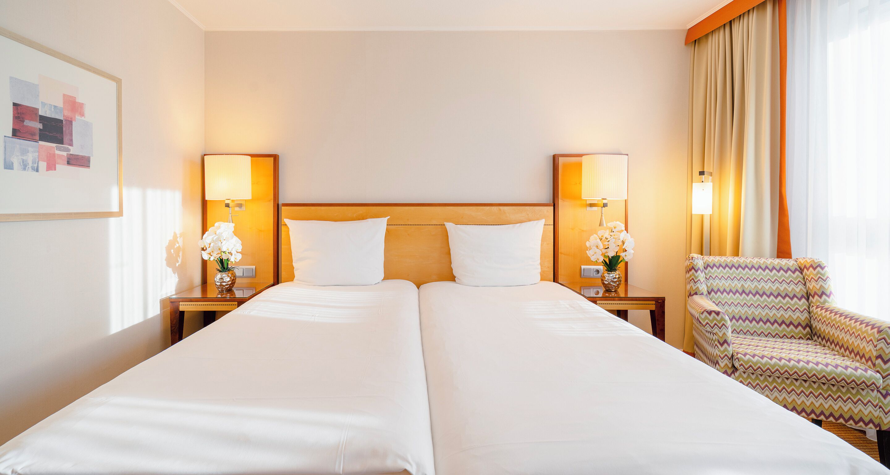 A hotel room with two beds, nightstands with lamps, a chair, and a wall-mounted artwork.