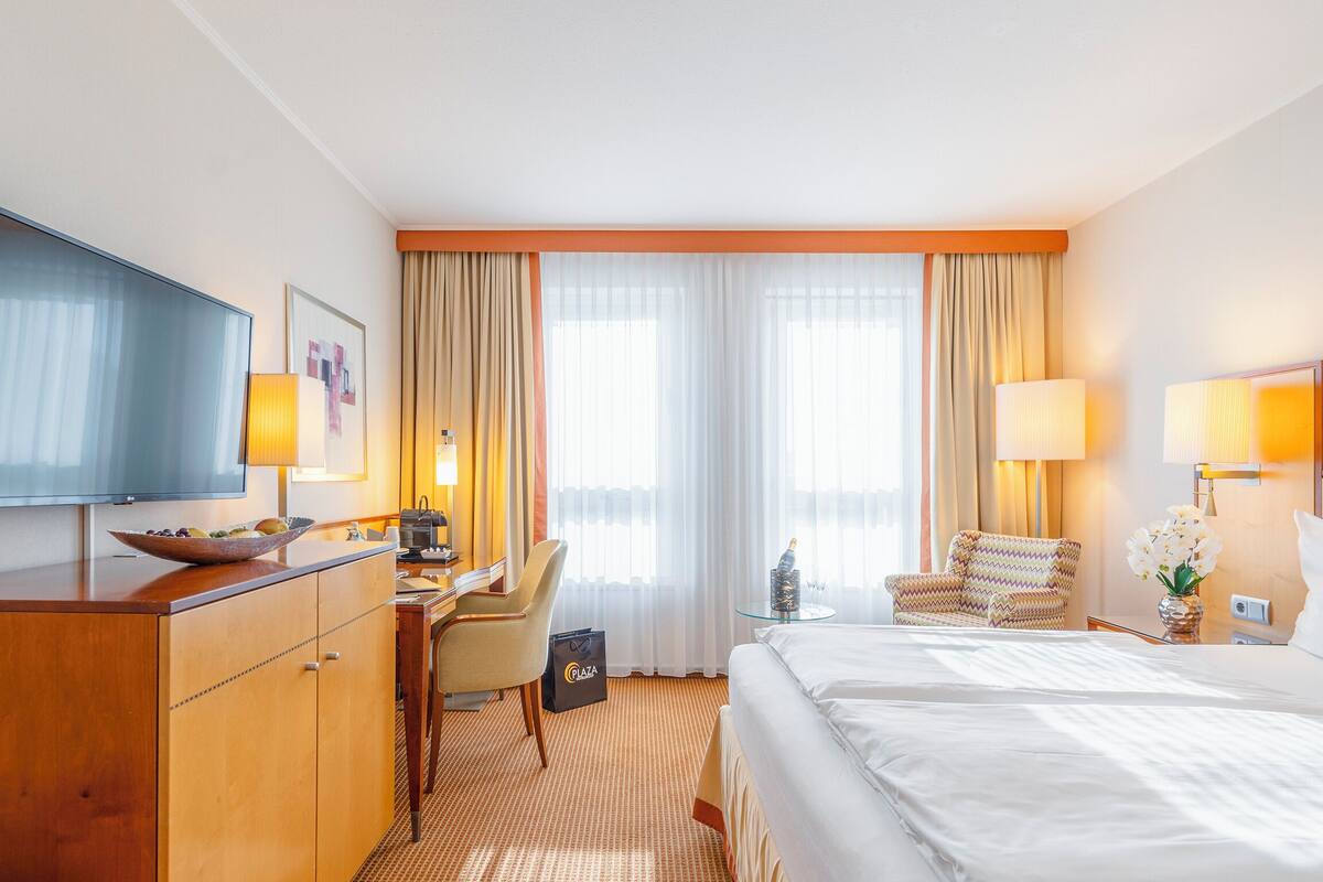Een hotelkamer met een flatscreen televisie, een houten bureau met een schaal fruit, een bed, een stoel en een klein tafeltje met een fles.