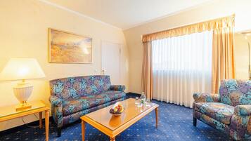 Ein Hotelzimmer mit einer gemusterten Couch, einem kleinen Tisch mit Lampe, einem Couchtisch mit Snacks und einem großen Fenster mit Vorhängen.
