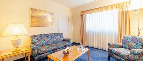 A hotel room with a patterned sofa, a small table with a lamp, a coffee table with snacks, and a large window with curtains.