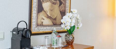 Una cafetera, un florero con forma de piña y orquídeas blancas, y una obra de arte enmarcada sobre una mesa de madera.