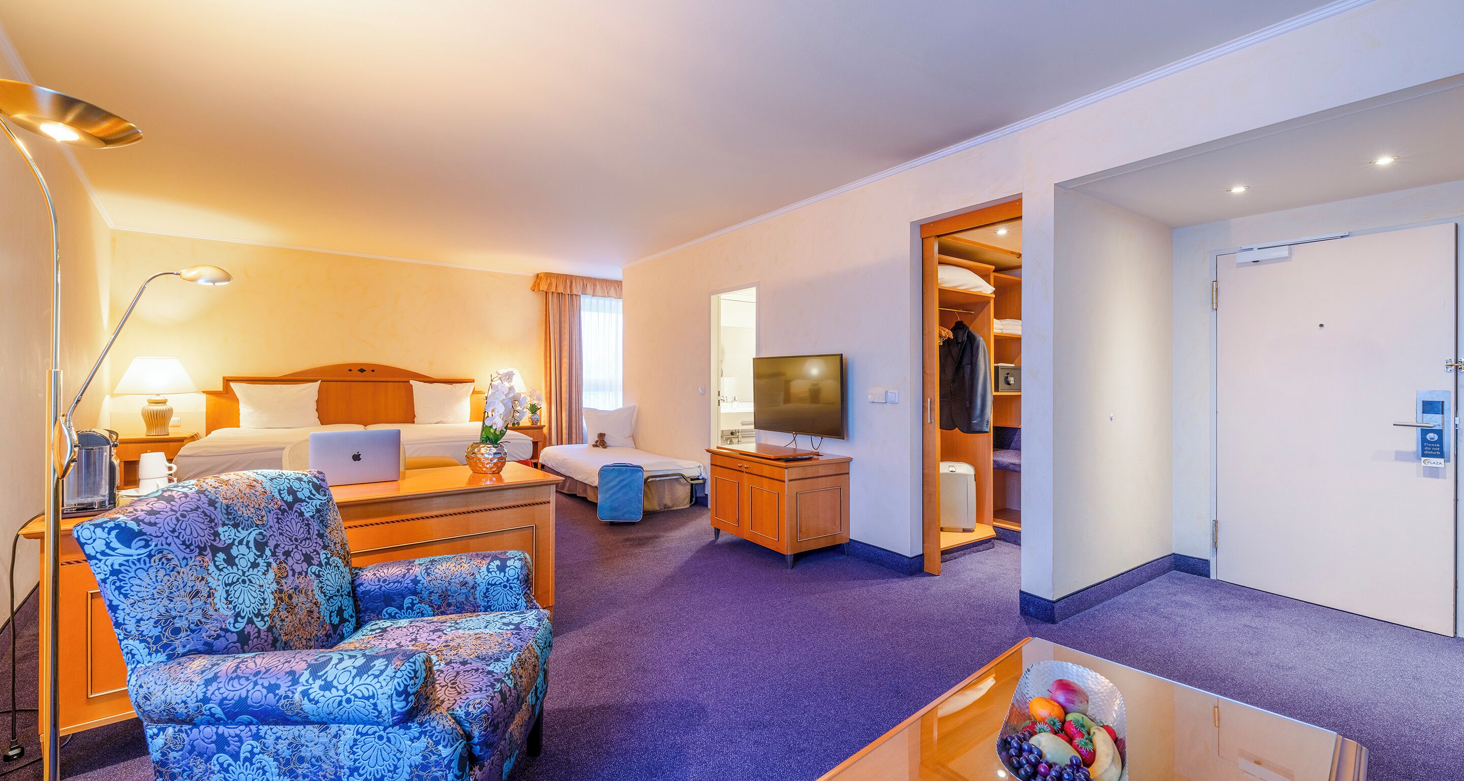 A hotel room with a bed, a desk with a laptop, a chair, a TV, and a wardrobe.