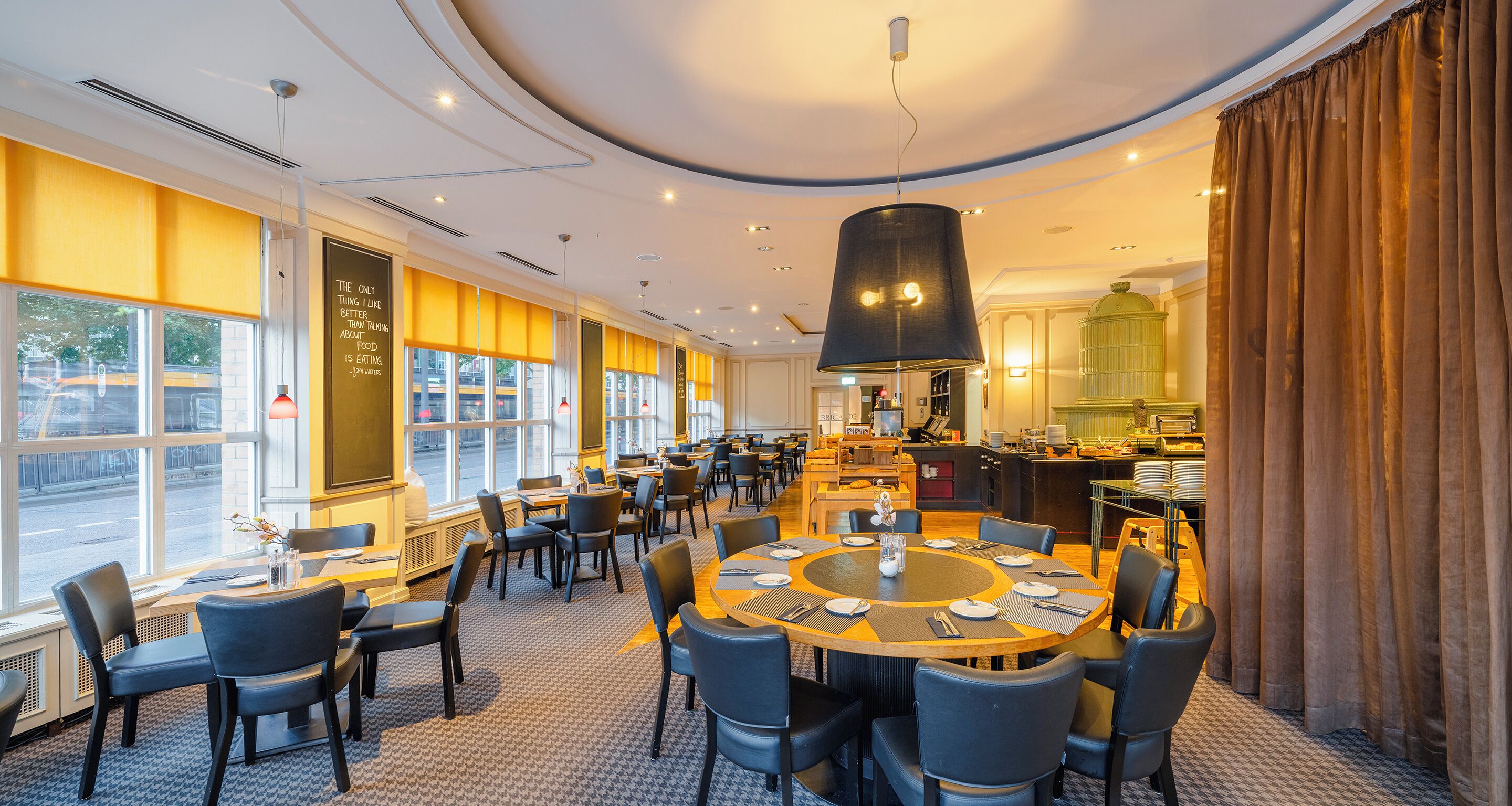 A spacious restaurant with a large round table, multiple dining tables, and chairs. There are large windows along one wall, a kitchen area, and a decorative birdcage.