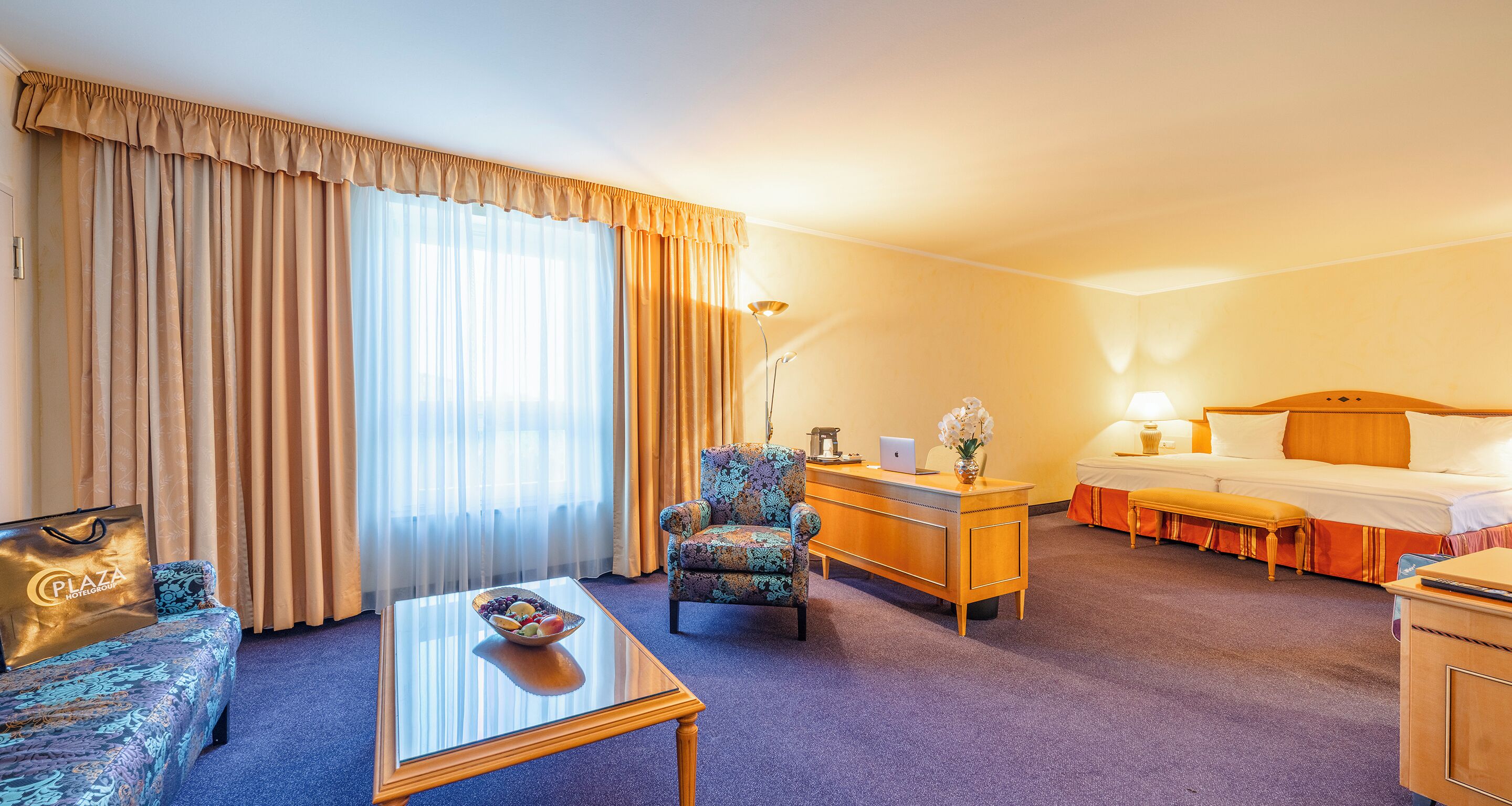 A hotel room with a sofa, a coffee table, a desk with a laptop, a bed, and a nightstand.