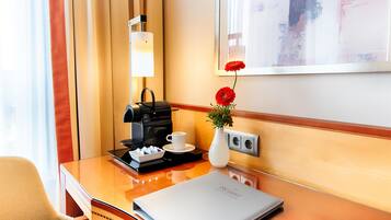 A hotel room with a wooden desk, a coffee maker, a white vase with red flowers, a framed artwork on the wall, and a window with curtains.