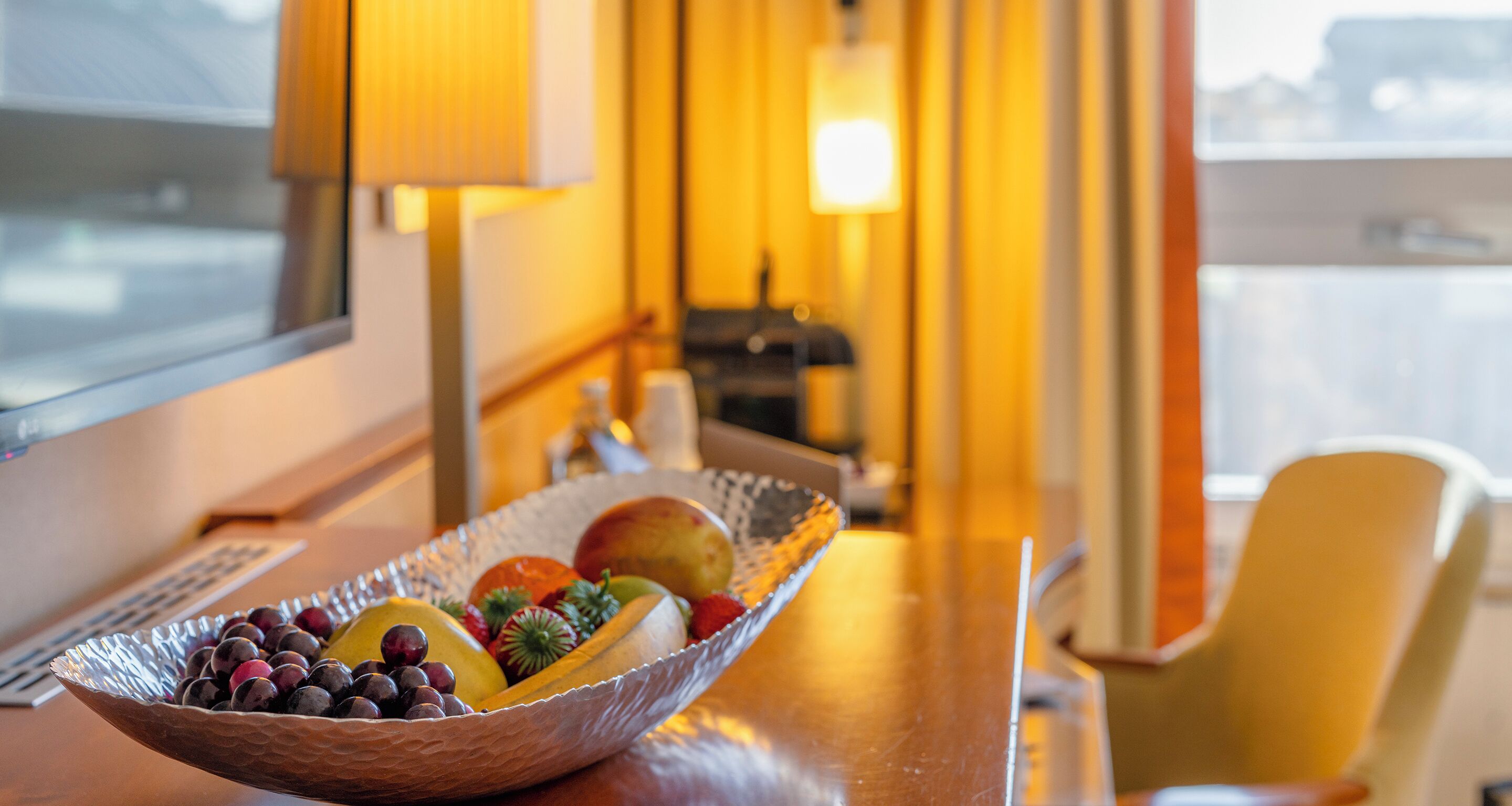 A fruit bowl on a wooden table with a chair and a lamp in the background.