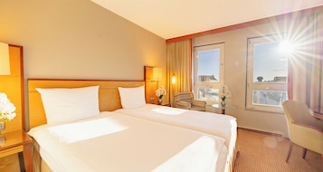 Habitación de hotel con dos camas, un espacio de estar con una silla, una mesita y una ventana con cortinas.