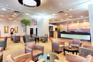 A spacious lobby with multiple seating areas, a large screen displaying a building, and a central column with a plant.