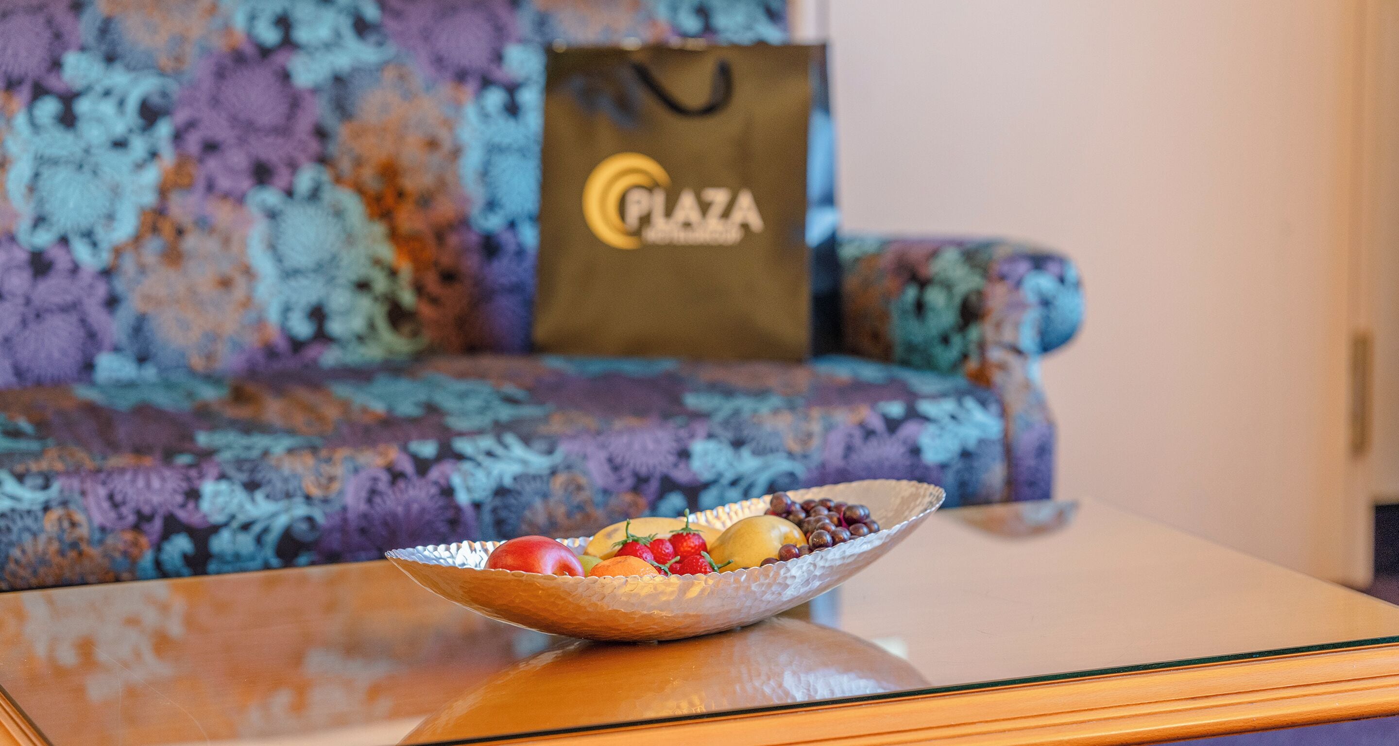A bowl of fruit on a glass table with a floral sofa and a 'Plaza' branded bag in the background.