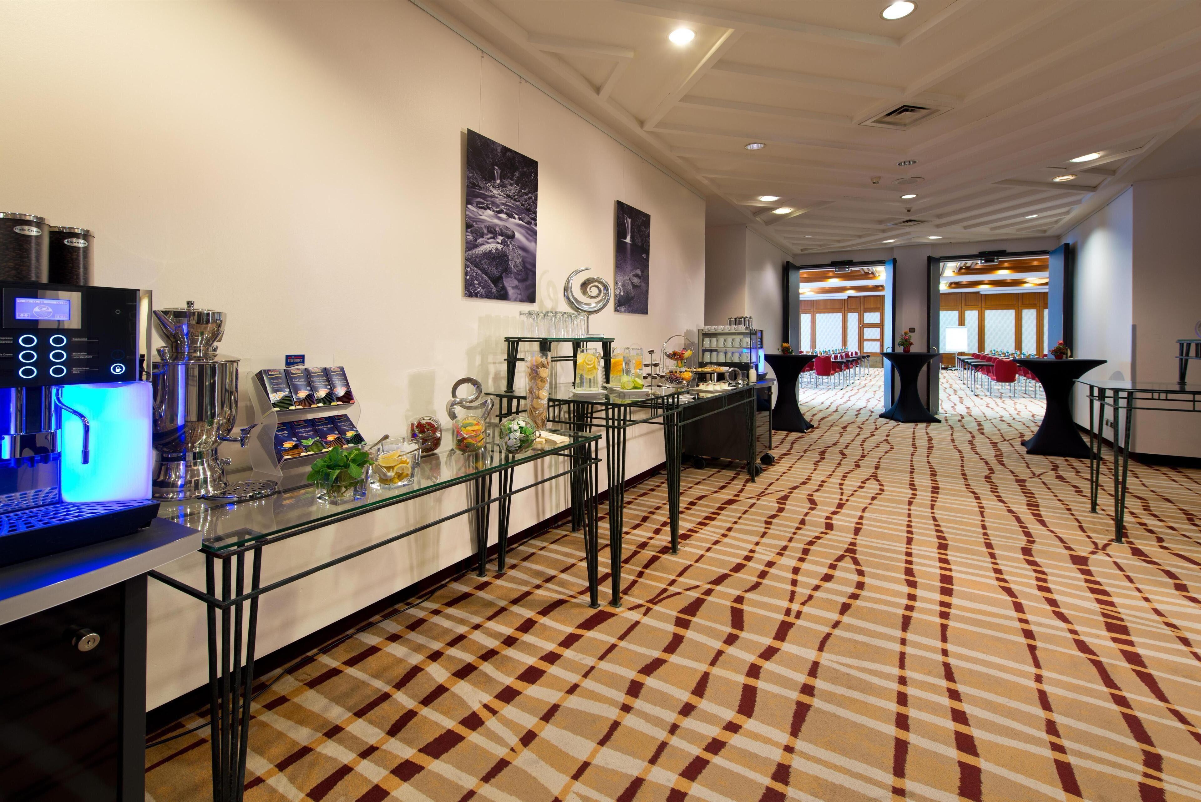 A hotel lobby with a check-in counter, a coffee machine, and a buffet area with various food items and drinks.