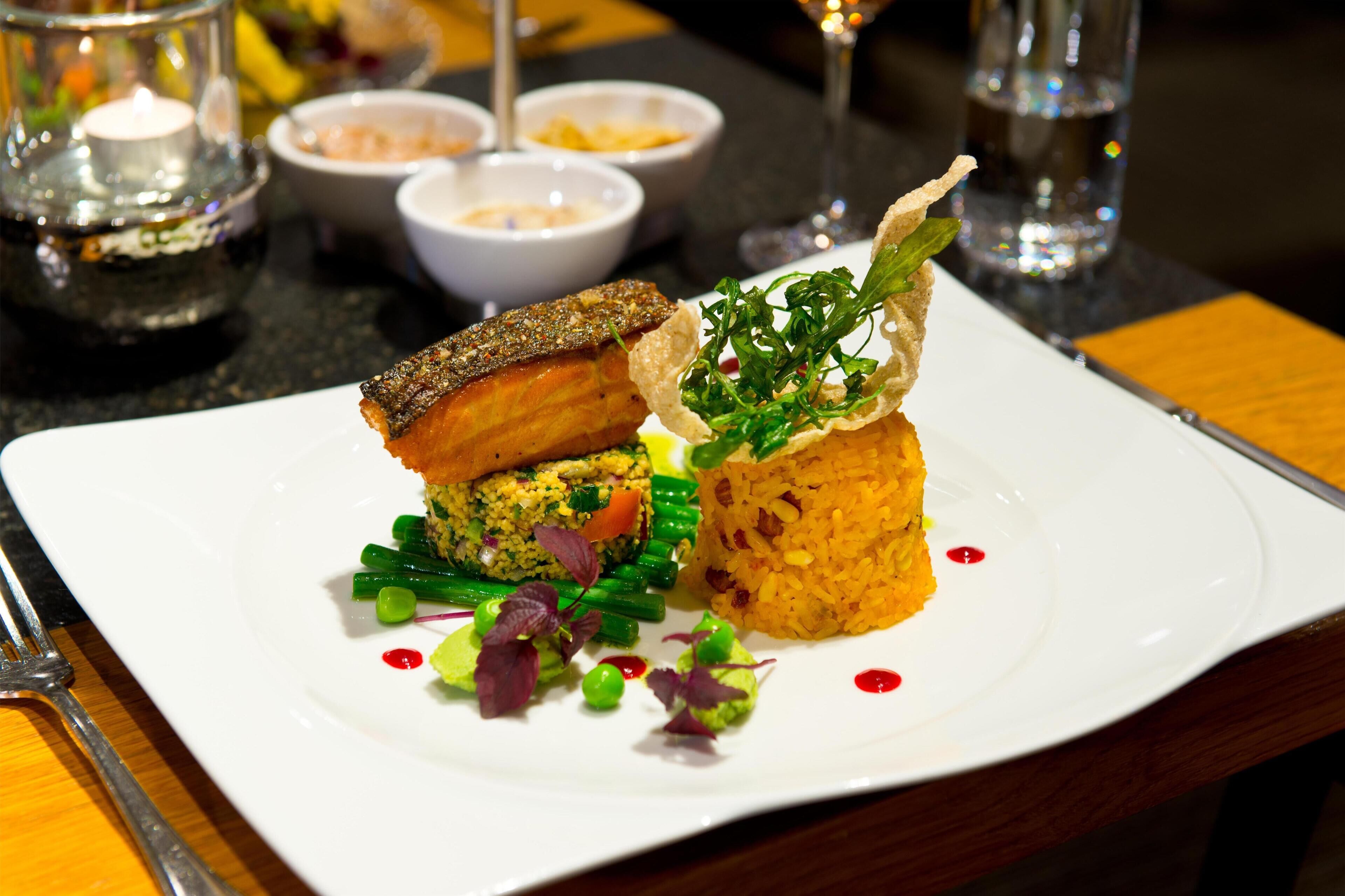 A white plate with a variety of food items including a piece of grilled meat, a vegetable patty, and a rice dish, accompanied by green beans and garnishes.