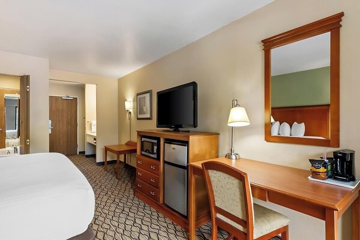 Habitación de hotel con cama, escritorio con lámpara, televisor, refrigerador y cafetera.