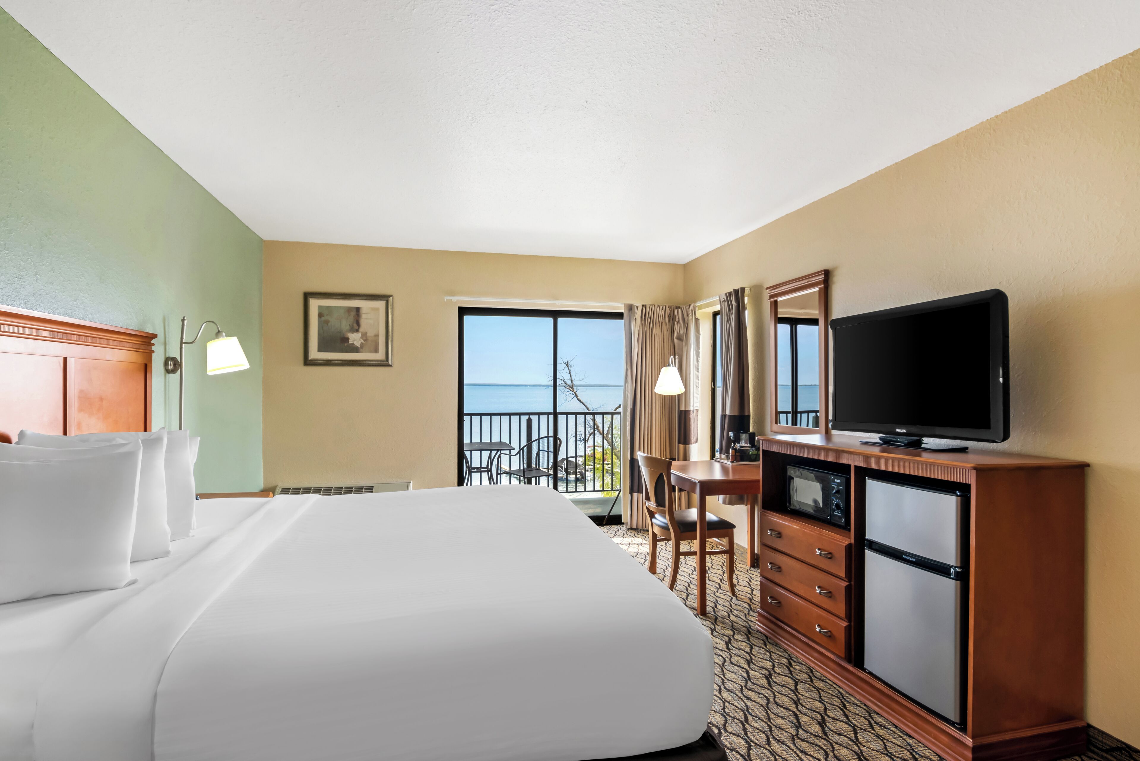 Une chambre d’hôtel avec un grand lit, un bureau avec une télévision, un réfrigérateur et une vue sur l’océan par la fenêtre.