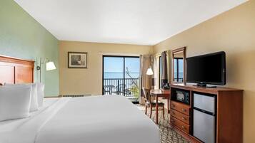 A hotel room with a large bed, a desk with a television, a refrigerator, and a view of the ocean through the window.