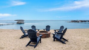 A beach with four Adirondack chairs arranged around a fire pit, overlooking a calm sea and a dock.