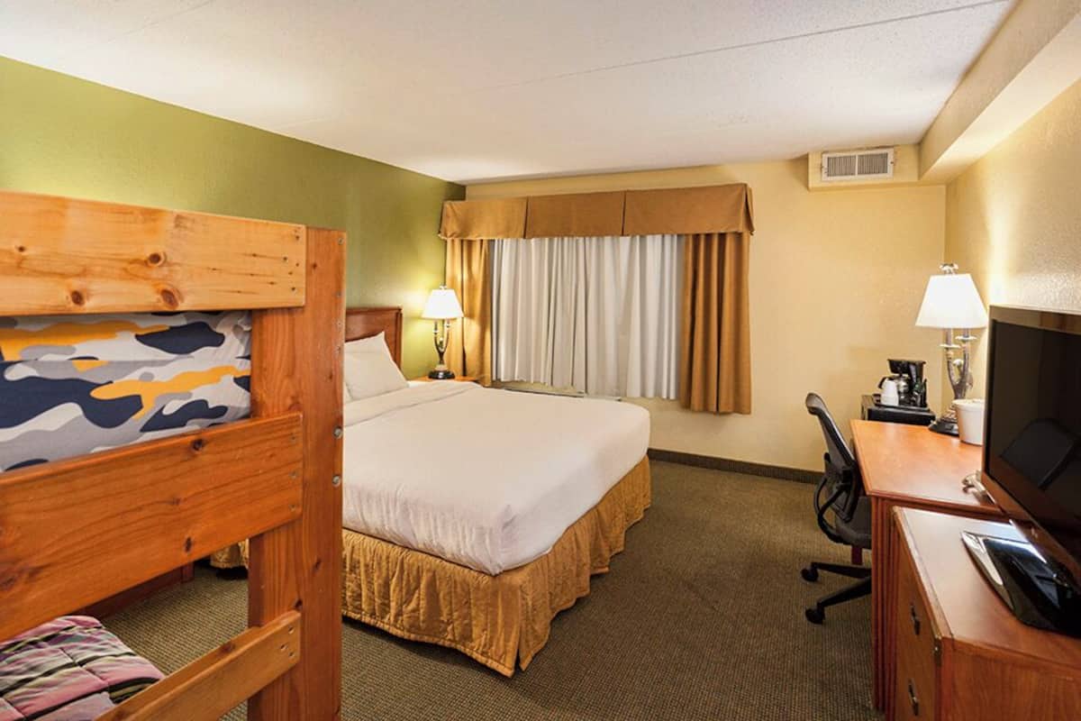 Une chambre d’hôtel avec un lit superposé, un lit simple, un bureau avec un ordinateur, une télévision et une fenêtre avec des rideaux.
