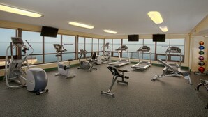 Ein Fitnessstudio mit unterschiedlichen Trainingsgeräten, großen Fenstern mit Meerblick und einer Decke mit Leuchtstofflampen.