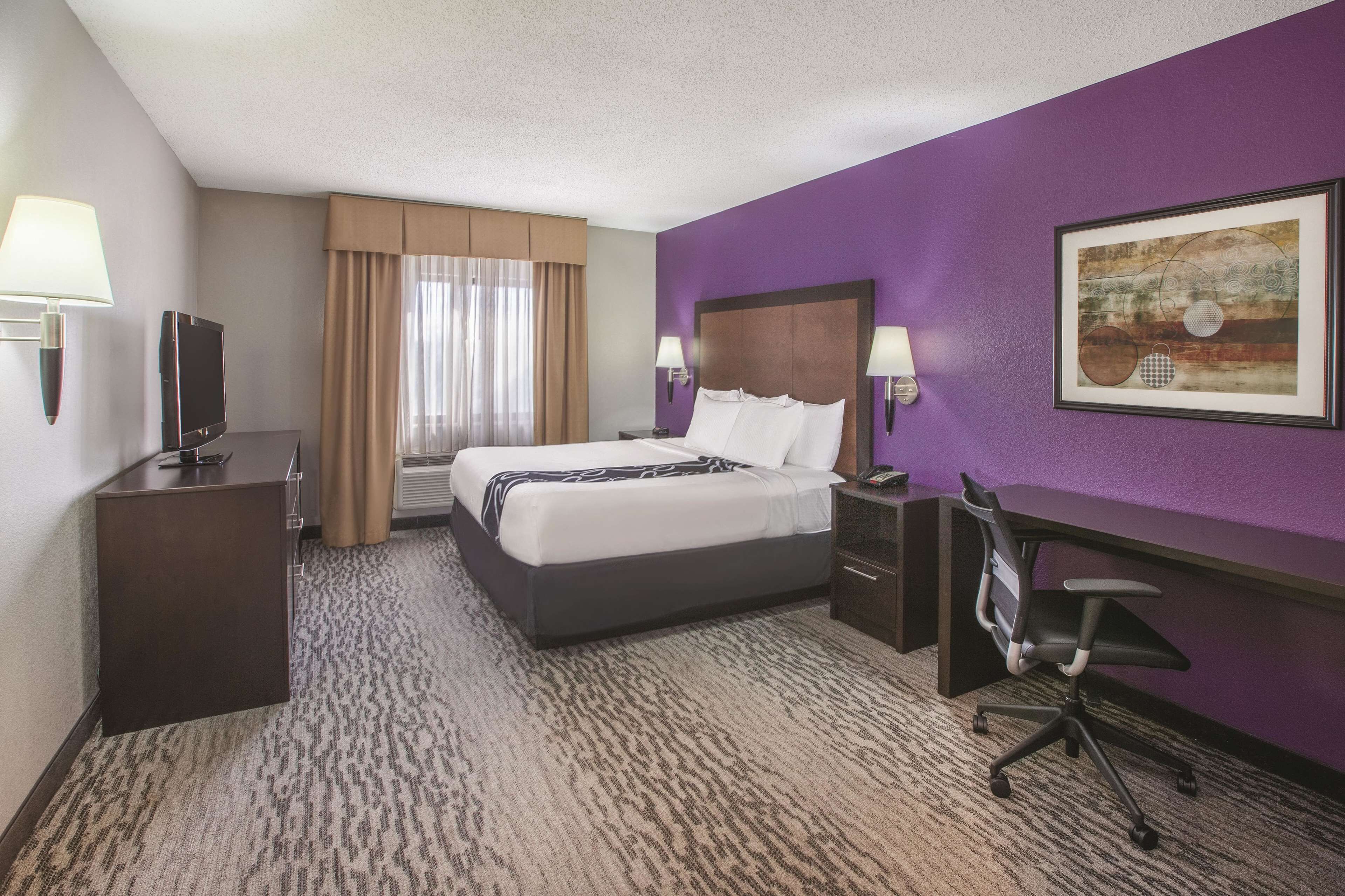 A hotel room with a bed, desk, chair, and a painting on the wall.