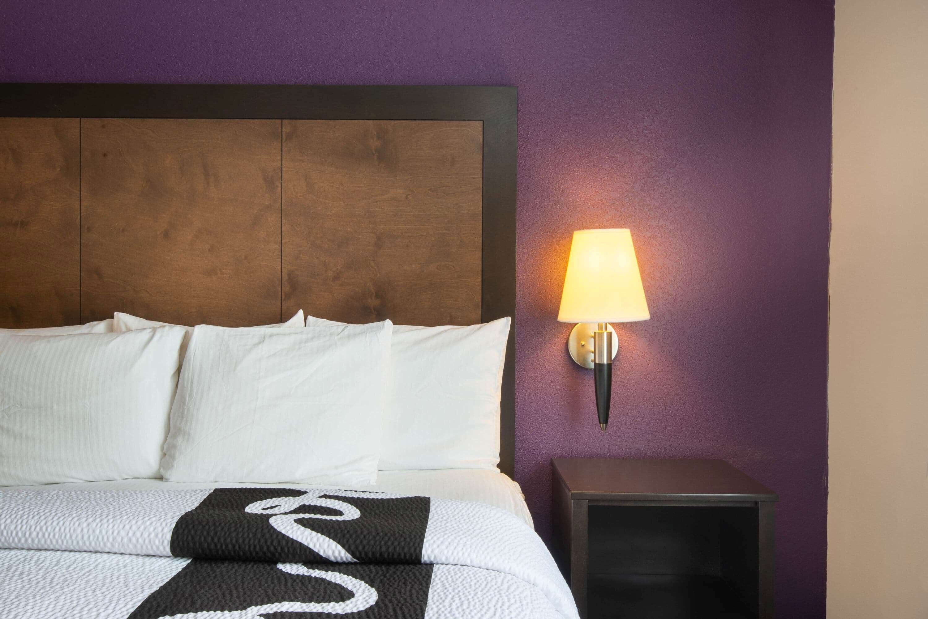 A neatly made bed with white linens and a black and white blanket, a wooden headboard, a nightstand with a lamp, and a purple wall.