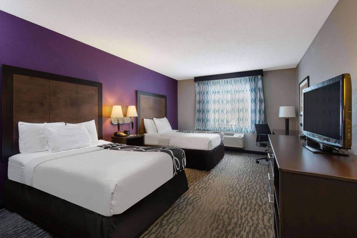 A hotel room with two beds, a desk, a television, and a window with patterned curtains.