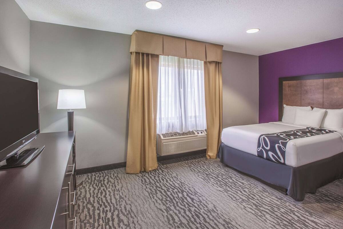A hotel room with a bed, a TV, a lamp, a window with curtains, and a carpeted floor.