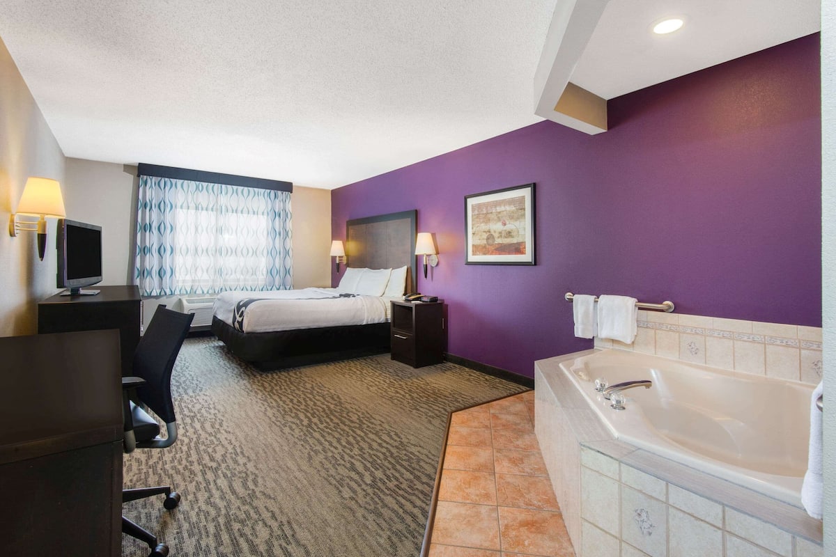 A hotel room with a bed, desk, chair, TV, lamps, and a bathtub.