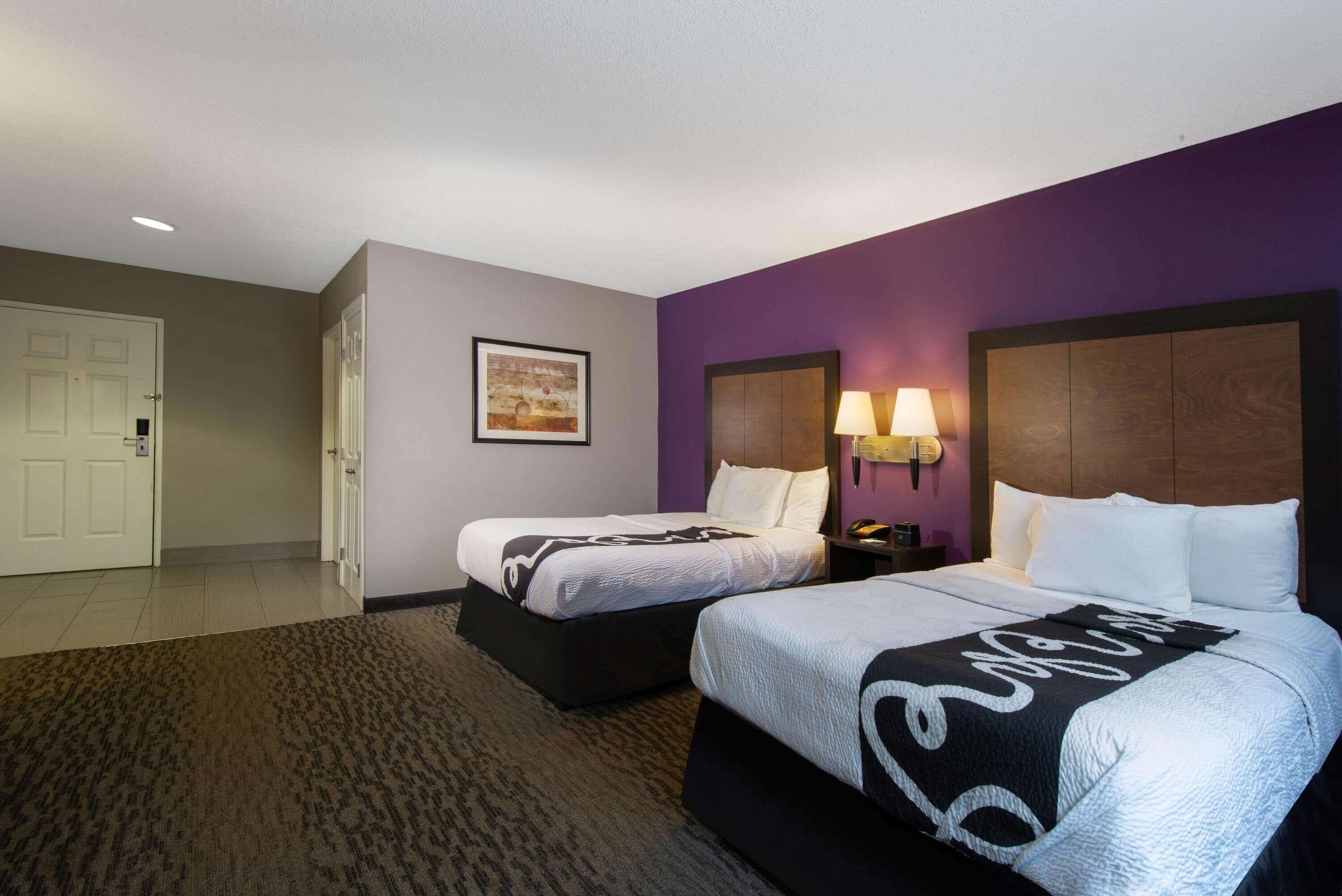 A hotel room with two beds, a purple accent wall, and a door to the left.
