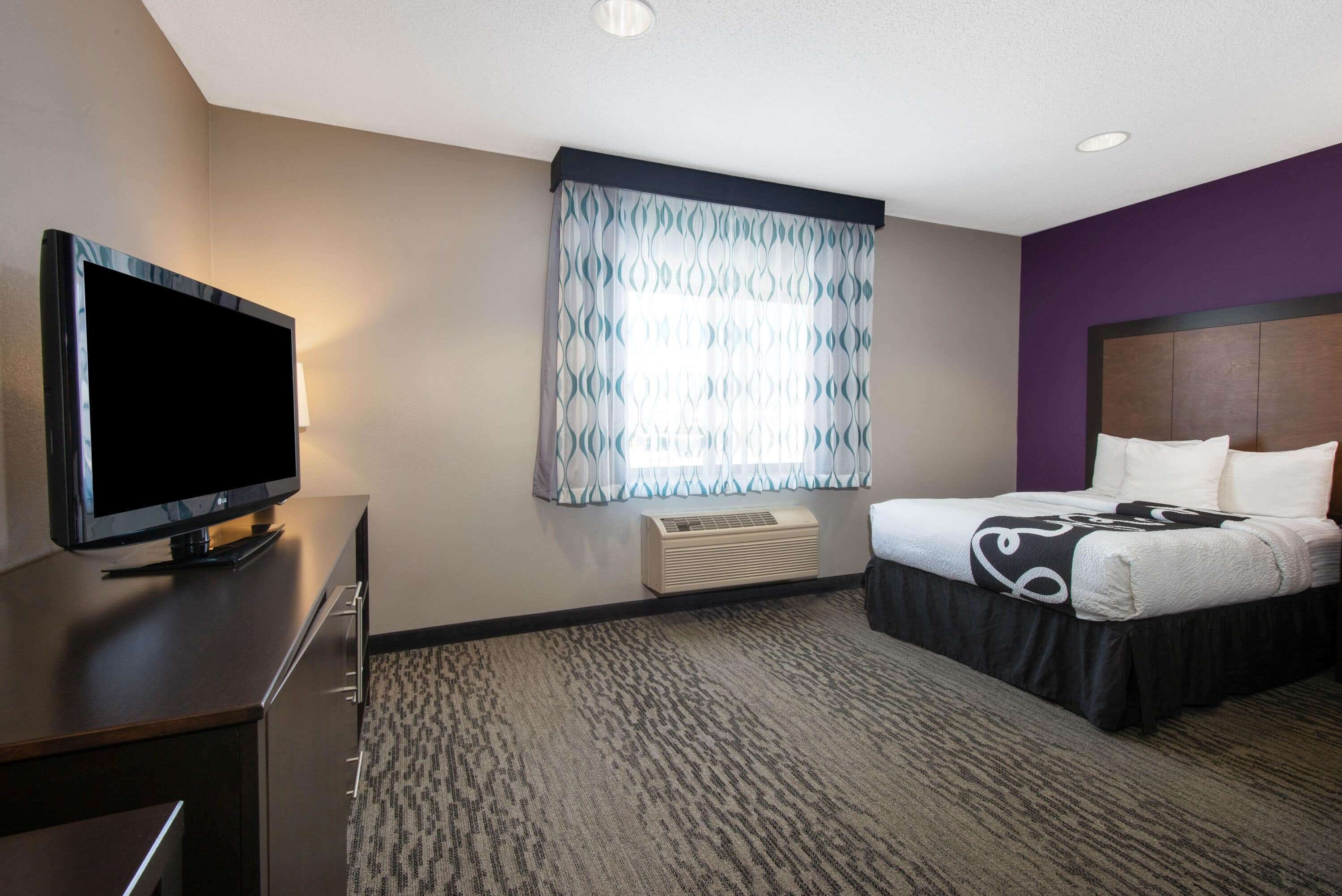 A hotel room with a bed, a TV on a dresser, a window with patterned curtains, and an air conditioning unit.