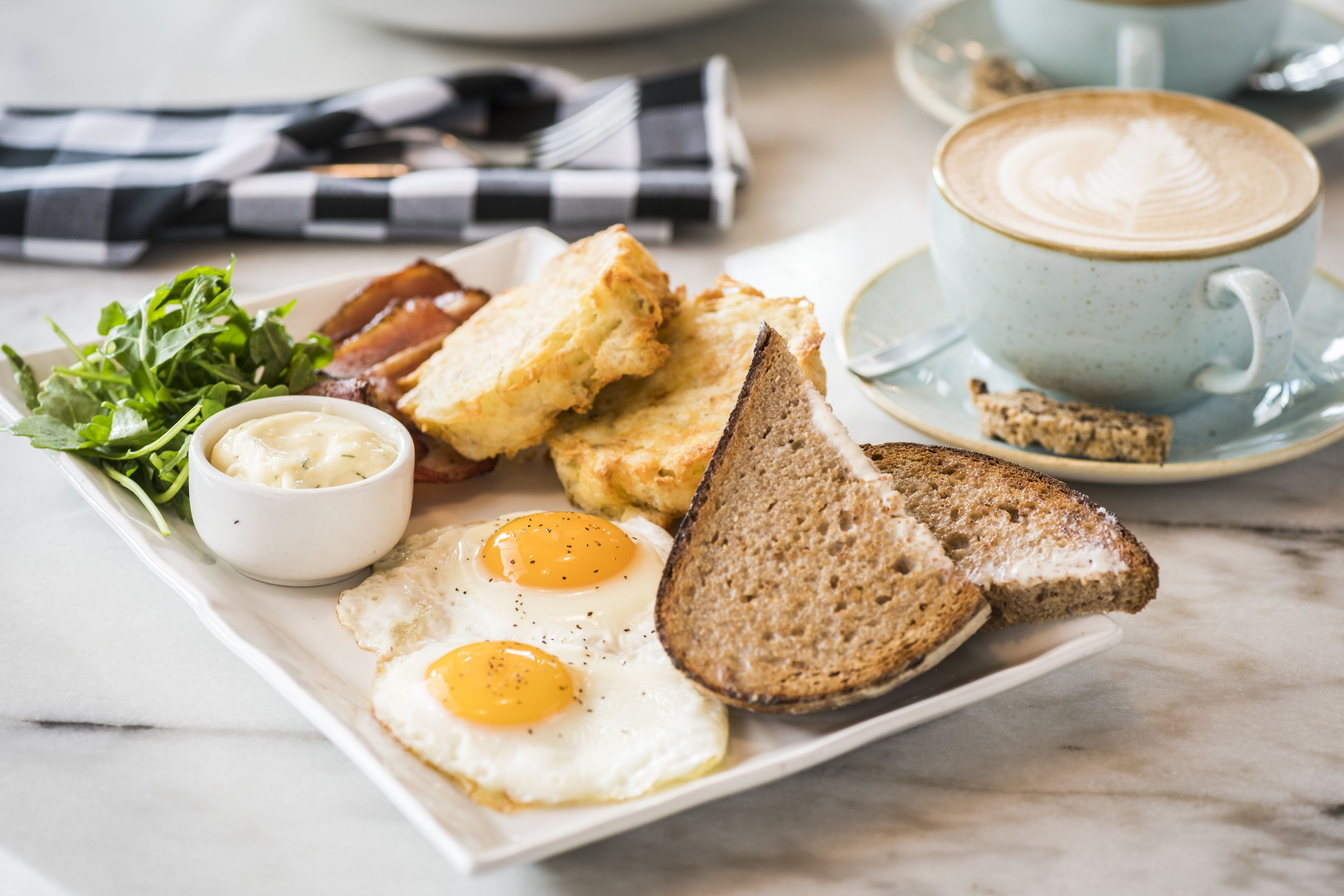 Une assiette de petit-déjeuner avec des œufs, des toasts, du bacon et des légumes verts, ainsi qu'une tasse de café sur une table.