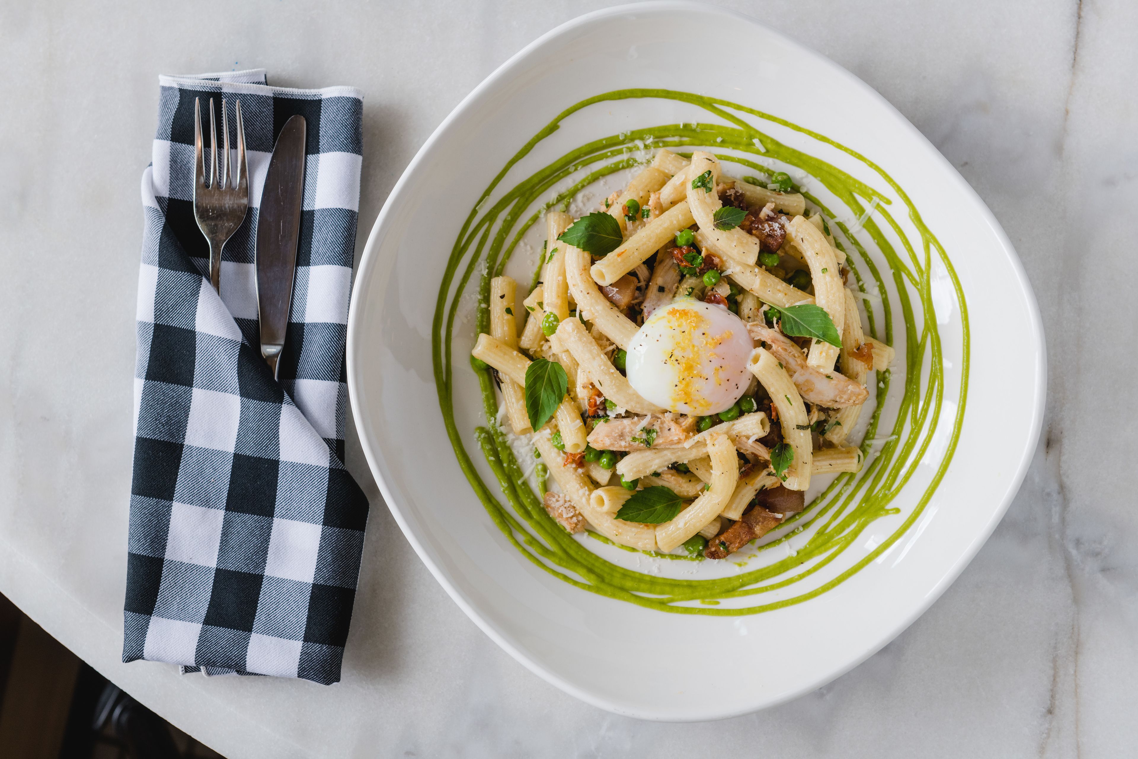 Plato blanco con pasta, un huevo pochado y salsa verde, acompañado de una servilleta de cuadros y cubiertos.