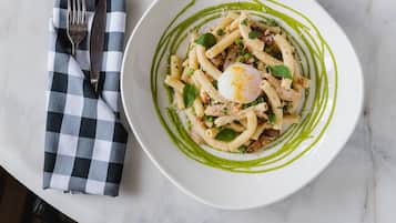 Plato blanco con pasta, un huevo pochado y salsa verde, acompañado de una servilleta de cuadros y cubiertos.